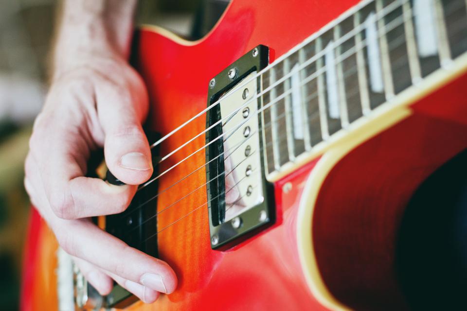 A guitarist's picking hand holding a pick over the strings of a red electric guitar